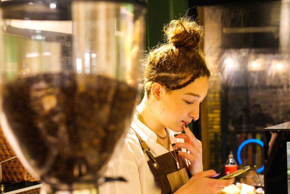 A focused young barista checks her phone while working in a cozy coffee shop setting.